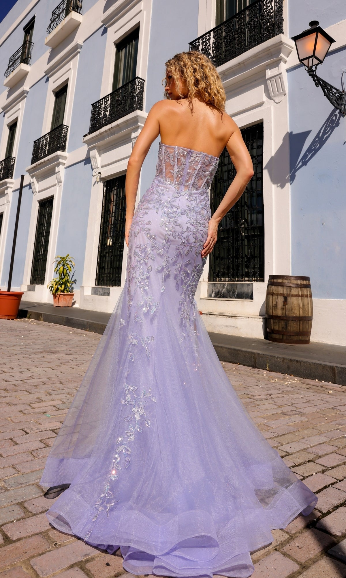A woman in the Sheer-Bodice Strapless Mermaid Prom Dress G1258 stands on a cobblestone street in front of a light blue building.